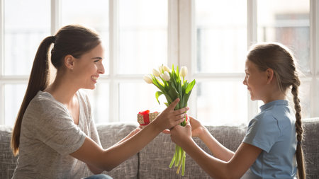 Caring little teen Caucasian daughter congratulate excited young mother with birthday anniversary. Loving small girl child greeting make surprise to happy mom, present flowers bouquet and gift.の写真素材
