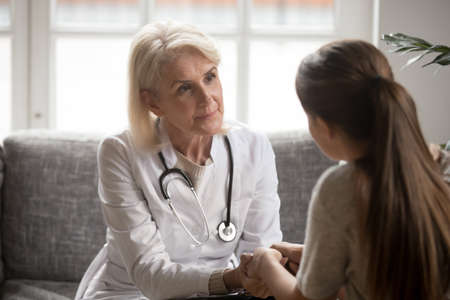 Attentive senior female doctor hold patient hands comfort support in difficult situation or illness. Caring woman GP or therapist feel supportive caress talk with client in hospital or clinic.の写真素材