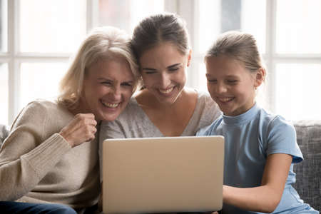 Happy three generations of Caucasian women look at laptop screen laugh talking on video call online....