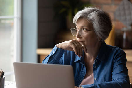 Old age and tech. Thoughtful aged latin female sit at desk work online by laptop from home office. Pensive lady retiree learn to use internet study computer ponder on next step distracted from screenの写真素材
