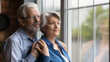 Looking in future. Loving senior man husband embrace aged wife from behind support encourage share feeling of beloved woman. Elderly spouses hug look at window meet twilight years together. Copy spaceの写真素材