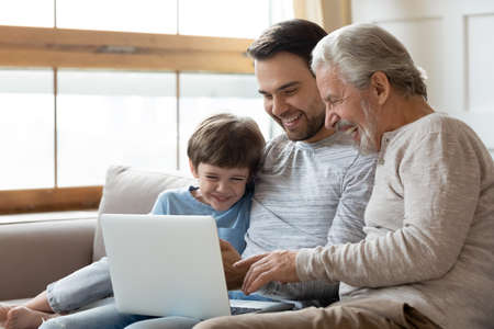 Overjoyed three generations of Caucasian men relax on couch look at laptop screen watch funny video...