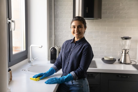 Portrait smiling Indian woman housewife wearing blue rubber gloves cleaning kitchen table surface,...