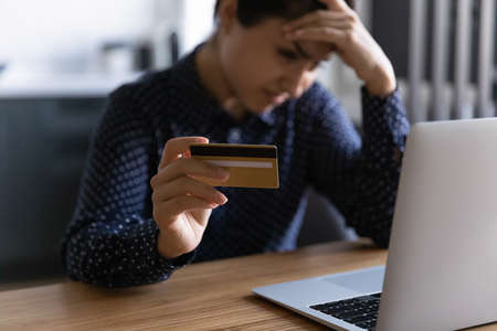 Close up worried Indian young woman holding credit card, having problem with money, unexpected debt, frustrated client touching forehead, using laptop, checking balance online, internet fraudの写真素材