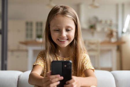 Smiling little Caucasian girl child hold smartphone have webcam online digital virtual event conference. Happy small 8s kid use cellphone talk speak on video call on gadget. Communication concept.の写真素材