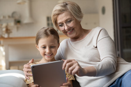 Smiling mature grandmother and small granddaughter look at tablet screen talk speak on video call online. Happy senior granny and little girl child have fun use modern pad device at home on weekend.の写真素材