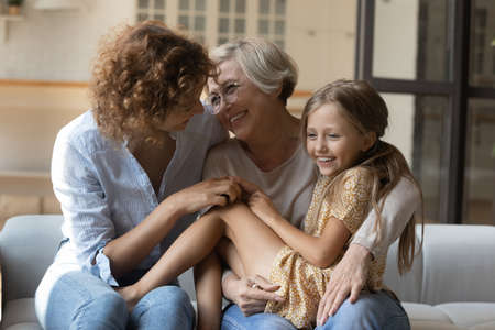 Overjoyed three generations of women relax on sofa at home have fun play together on weekend. Smiling Caucasian little 9s girl child enjoy playful family time with young mom and senior grandmother.の写真素材