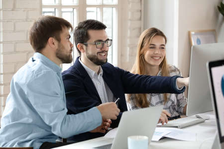 Smiling multiracial colleagues gather at workplace look at PC screen brainstorm cooperate together. Happy diverse businesspeople look at computer screen discuss business project. Teamwork concept.の写真素材