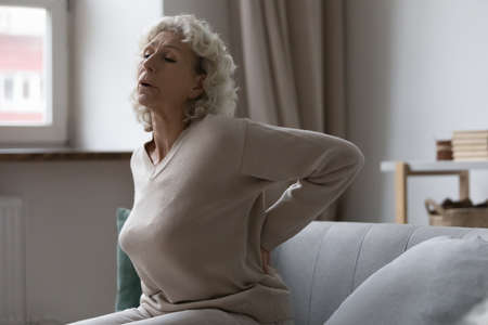 Frustrated senior woman suffering from rheumatism, radiculitis when trying to stand up at home. Unhappy mature elder lady feeling lumber pain, painful muscles stiffness, holding lower back.の写真素材