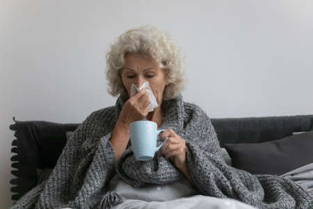 Sick mature senior woman suffering from flu, cold, respiratory infection. Elderly 60s lady wrapped...