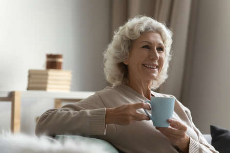 Happy elderly lady enjoying cup of hot beverage at home, looking away, smiling. Mature 60s woman...