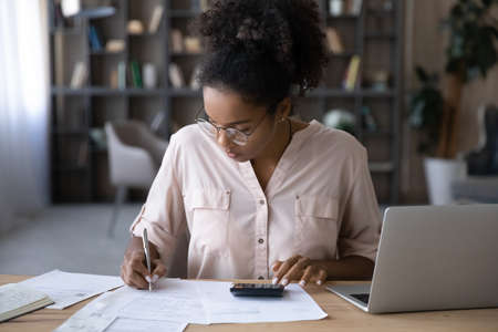 Serious young biracial woman sit at desk manage budget calculate on machine pay bills taxes online on laptop. Focused African American female count expenses expenditures on calculator. Save concept.の写真素材