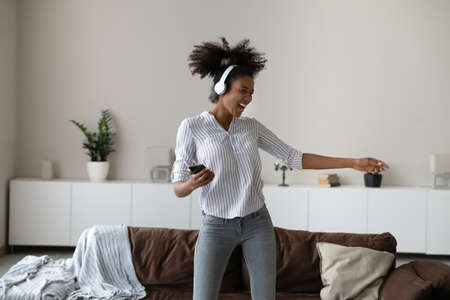 Excited young African American woman in modern headphones listen to music on smartphone dance at home. Happy biracial female in earphones have fun enjoy good quality sound on cellphone gadget.の写真素材