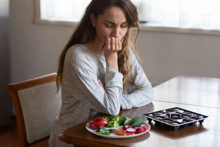 Unhappy young Latin woman look at chocolates and vegetables face temptation suffer from eating disorder. Millennial female think of healthy food choice, diet. Wellbeing, weight loss concept.の写真素材