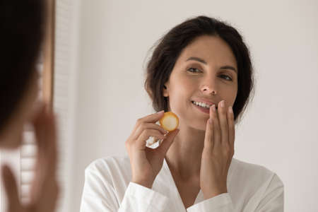 Head shot mirror reflection beautiful woman applying moisturizing lip balm or mask on dry lips,...