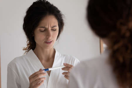 Close up mirror reflection of unhappy woman holding pregnancy test, frustrated young female in bathrobe dissatisfied by result, standing in bathroom, unwanted pregnancy or health problem, infertilityの写真素材