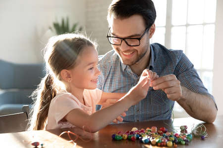 Playful young Caucasian father and little happy daughter have fun making bracelets accessories at home together. Loving dad and small girl child engaged in funny hobby creative activity string beads.の写真素材
