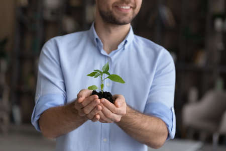 Crop close up of smiling young Caucasian male leader or boss hold sprout or seedling in hands planning. Happy millennial businessman with soil and green plant. Development, startup launch concept.の写真素材