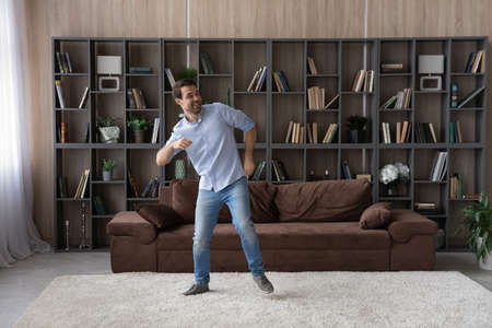 Overjoyed young Caucasian man dance in cozy new renovated living room have fun on weekend at home. Happy millennial male renter or tenant celebrate moving relocation to own house or apartment.の写真素材