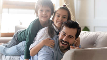 Portrait of smiling young Caucasian single dad and two small kids lying on sofa have fun feeling playful. Happy loving father and little children relax rest on couch at home on family weekend.の写真素材