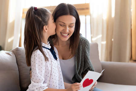 Caring small teen girl child congratulate happy young Caucasian mother with birthday anniversary....