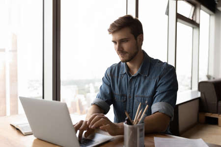Pensive young Caucasian man work online on laptop in home office, browse wireless internet on gadget. Millennial male employee busy consulting client or customer on computer at workplace.の写真素材