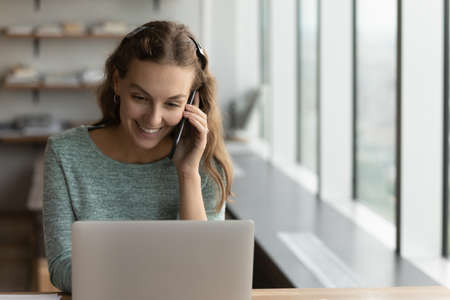 Happy young Caucasian businesswoman work on laptop in office talk speak on cellphone call with...