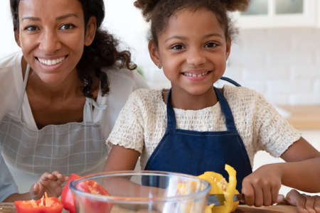 Portrait of happy mom and daughter wearing aprons, cooking in kitchen together. Sweet preschooler girl helping her mum to male salad for dinner, learn to slice fresh vegetables. Cropped head shotの写真素材