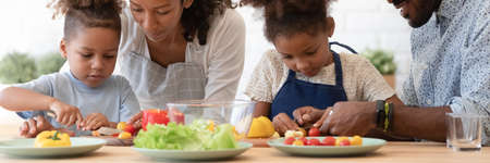 Happy African American parents teaching sibling kids to cook, slicing fresh vegetables, peppers, tomatoes for salad on kitchen table or counter. Family healthy eating, organic food concept. Bannerの写真素材