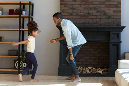 Happy African American dad teaching daughter girl to dance at home. Active daddy and kid exercising together, warming up in living room, enjoying home activities, listening to music, having funの写真素材