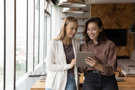 Smiling young female colleagues look at pad screen talk speak on video call at office meeting. Happy women employees use work together on tablet, have webcam zoom digital virtual event on gadget.の写真素材