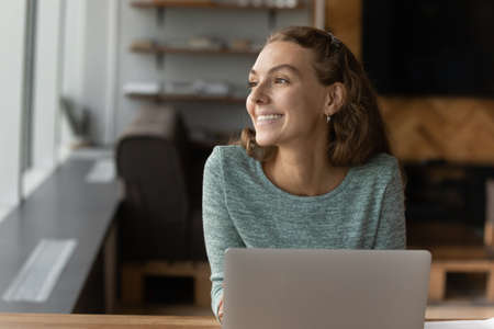 Overjoyed millennial female employee distracted from computer work look in window distance thinking or dreaming. Happy young Caucasian woman use laptop imagine or visualize. Business vision concept.の写真素材