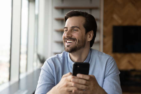 Overjoyed millennial man use hold modern smartphone look in distance dreaming thinking. Happy young Caucasian male have fun laugh browsing texting on cellphone. Communication, technology concept.の写真素材