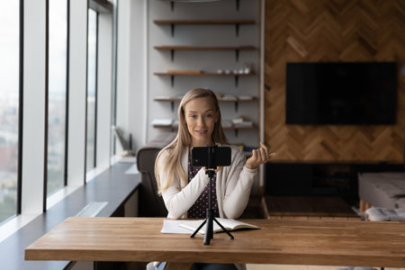 Young Caucasian woman sit in home office record video live broadcast on smartphone camera....
