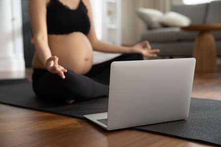 Crop close up of sporty pregnant woman sit on mat meditate practice yoga with online class on computer at home. Active female follow healthy lifestyle during pregnancy have webcam training on laptop.の写真素材