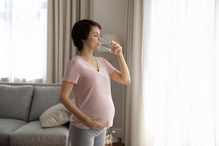 Young Caucasian pregnant woman drink clean clear still mineral water from glass feel dehydrated...