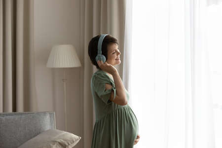 Smiling young Caucasian pregnant woman wear headphones listen to music enjoy maternity leave. Happy millennial female with pregnancy in earphones enjoy good quality sound in modern gadget.の写真素材