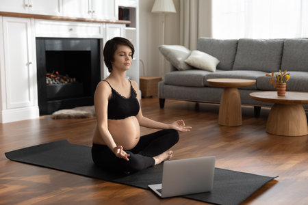 Calm young Caucasian pregnant woman sit on mat in living room practice yoga with webcam online lesson on laptop. Millennial female follow healthy lifestyle during pregnancy, have virtual training.の写真素材