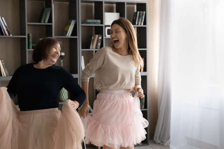 Excited mature Caucasian mother and grownup daughter wear ballroom princess skirts dance together at home. Smiling elderly mom and adult girl child have fun moving to rhythm music relaxing.の写真素材