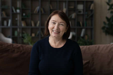 Headshot portrait of smiling modern elderly 60s Caucasian woman sit on sofa ta home talk speak on...