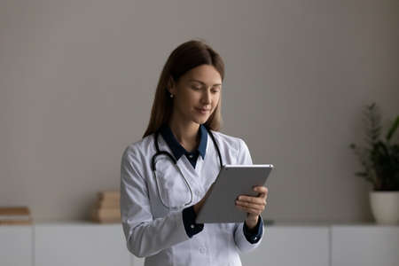 Focused female doctor in white medical uniform use modern tablet have webcam digital virtual conference. Young Caucasian woman nurse or GP talk on video call on pad device in hospital.の写真素材