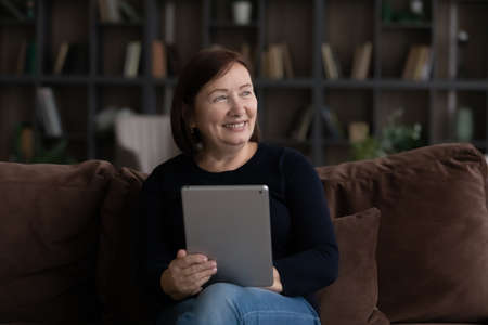 Smiling mature Caucasian woman hold use pad device look in distance thinking or planning. Happy...