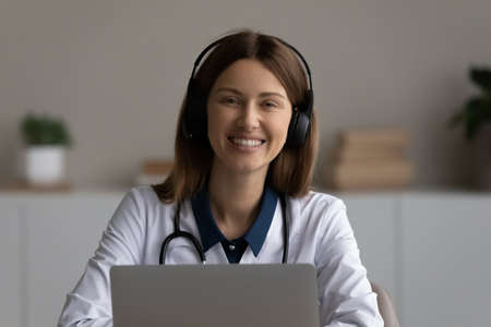 Portrait of smiling young Caucasian female nurse or GP in headphones talk on video call on laptop in...