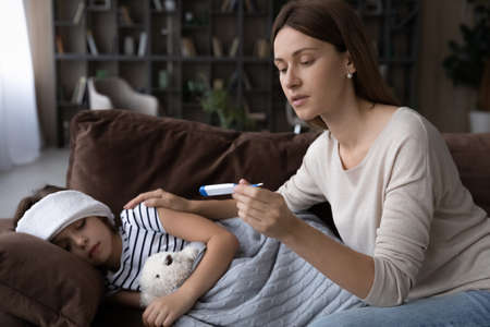 Worried young mother take care of sick small daughter feel unhealthy, check measure high temperature with thermometer. Anxious mom cure little child ill with flu or covid-19 virus. Healthcare concept.の写真素材