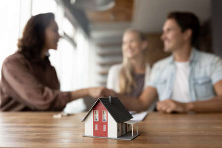 Crop close up of cardboard mock-up house model on table in real estate agent or broker office. Couple buyers shake hand of realtor on background. Spouses buy home make deal at meeting. Rental concept.の写真素材