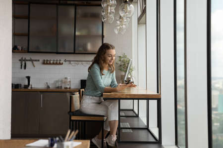 Young Caucasian female sit at desk in kitchen in office text or message online on cellphone device. Millennial woman relax on work break browse internet on smartphone gadget. Technology concept.の写真素材
