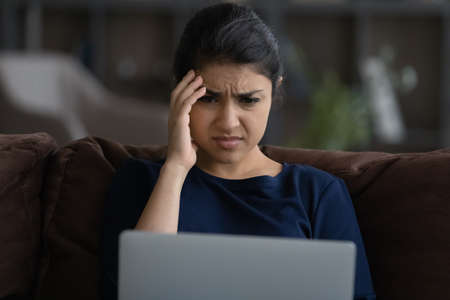 Close up unhappy Indian woman looking at laptop screen, reading bad news in message or social network, sitting on couch at home, worried businesswoman freelancer received dismissal notificationの写真素材