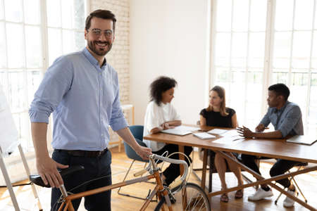 Happy young male manager in eyeglasses holding bicycle, looking at camera, enjoying healthy sporty...