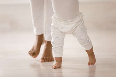 Crop close up of little African American baby toddler learn walking at home holding mom hands....
