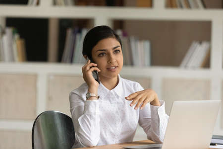 Close up smiling Indian businesswoman talking on phone, sitting at work desk with laptop in office,...